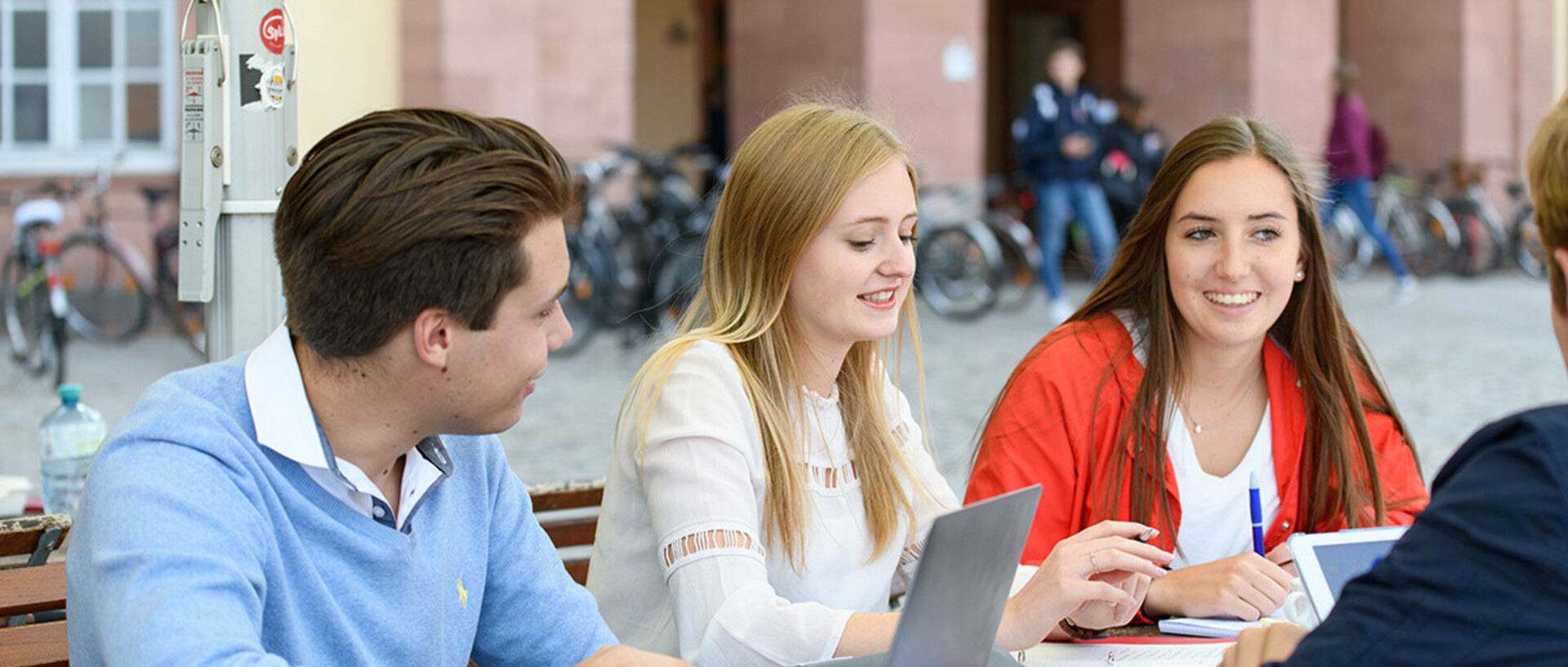 Studierende sitzen mit ihren Laptop draußen an einem Tisch. Im Hintergrund das Schloss